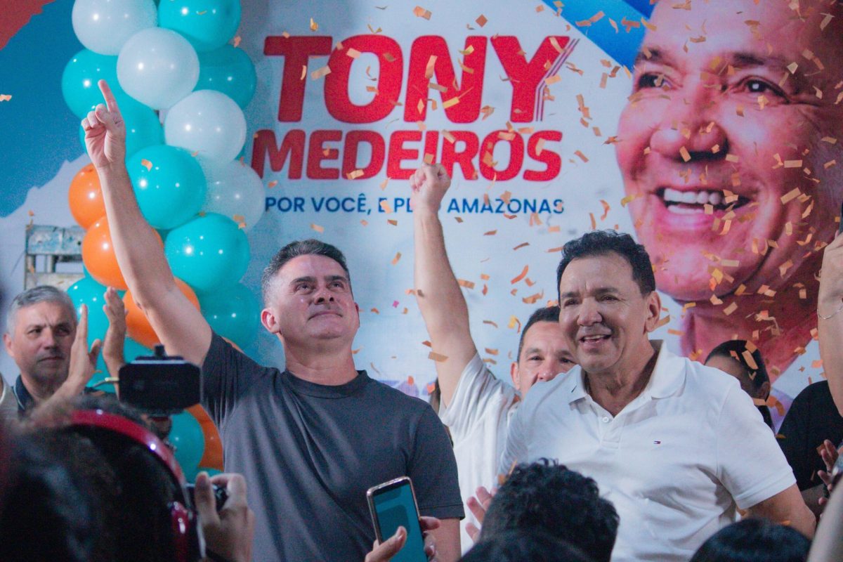 Dois homens levantam os braços durante evento político com público e painel de campanha ao fundo em Manaus.