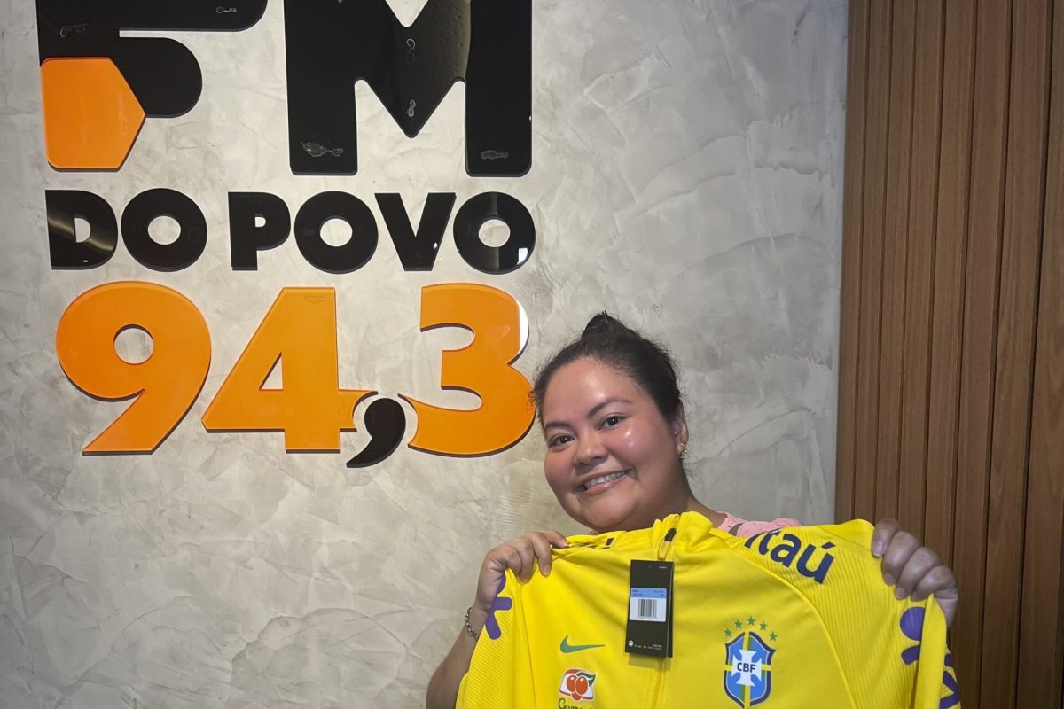 Neucilene Bentes Gato posa na sede da FM do Povo 94,3 segurando o agasalho oficial da Seleção Brasileira recebido como prêmio de uma promoção especial da Copa do Mundo de 2026.